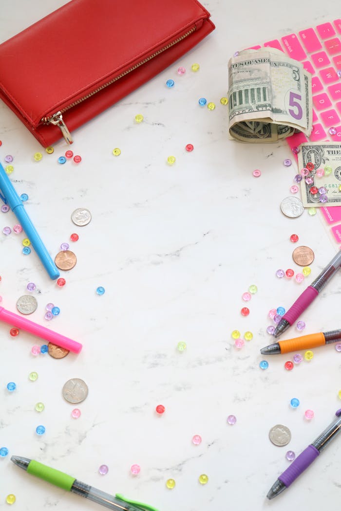 Flat lay of a creative workspace featuring currency, stationery, and colorful sequins on a marble surface.